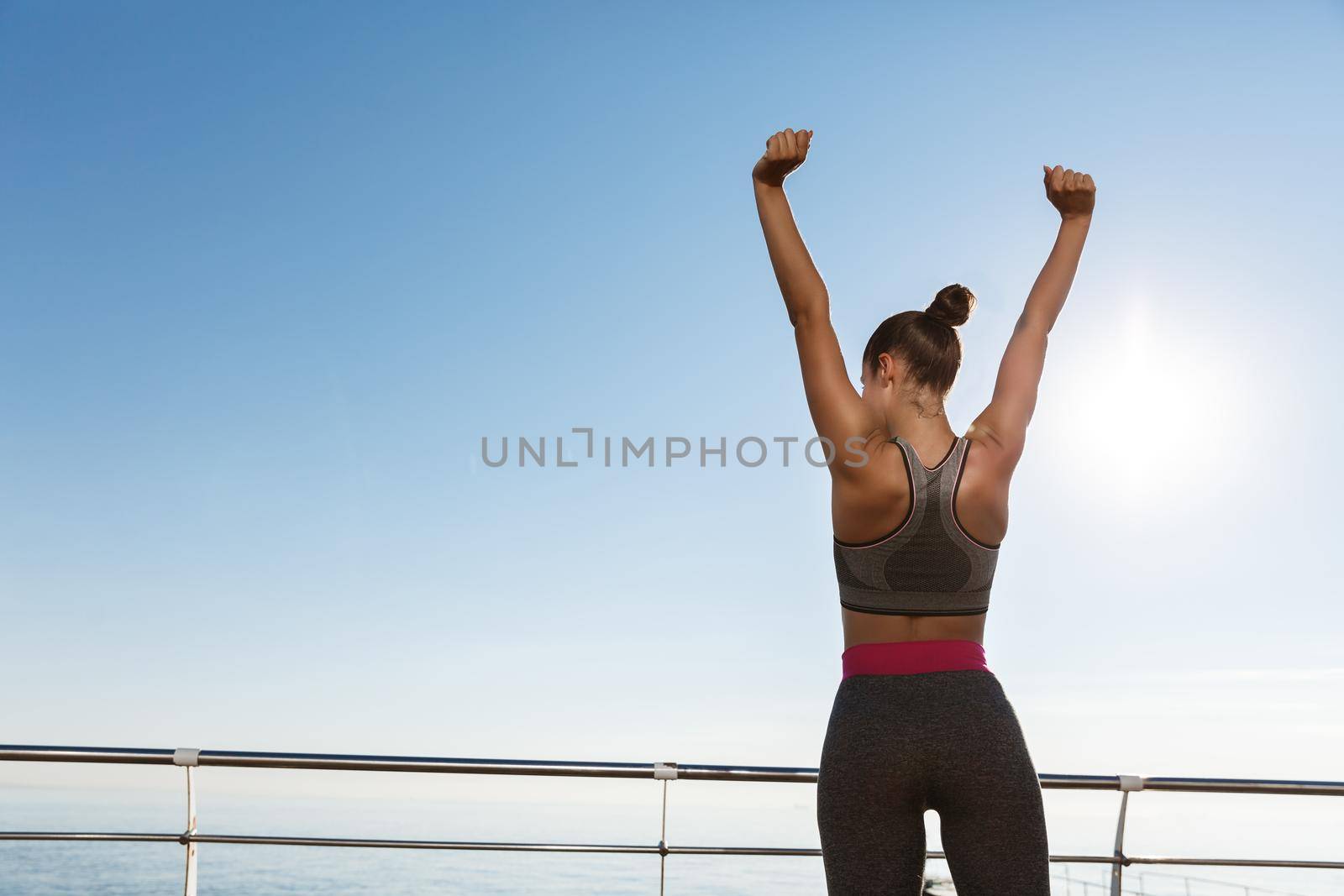 Rear view of happy triumphing sportswoman standing on a pier, raising hands up like champion, achieve goal during training session outdoors.