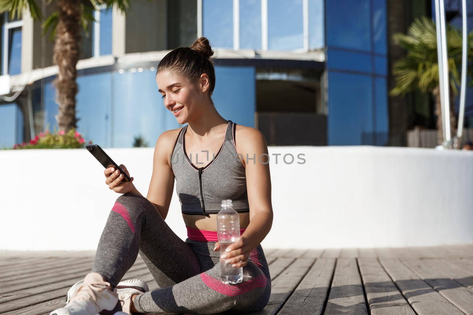 Outdoor shot of happy attractive sportswoman sitting on floor after workout, drinking water and looking at smartphone.