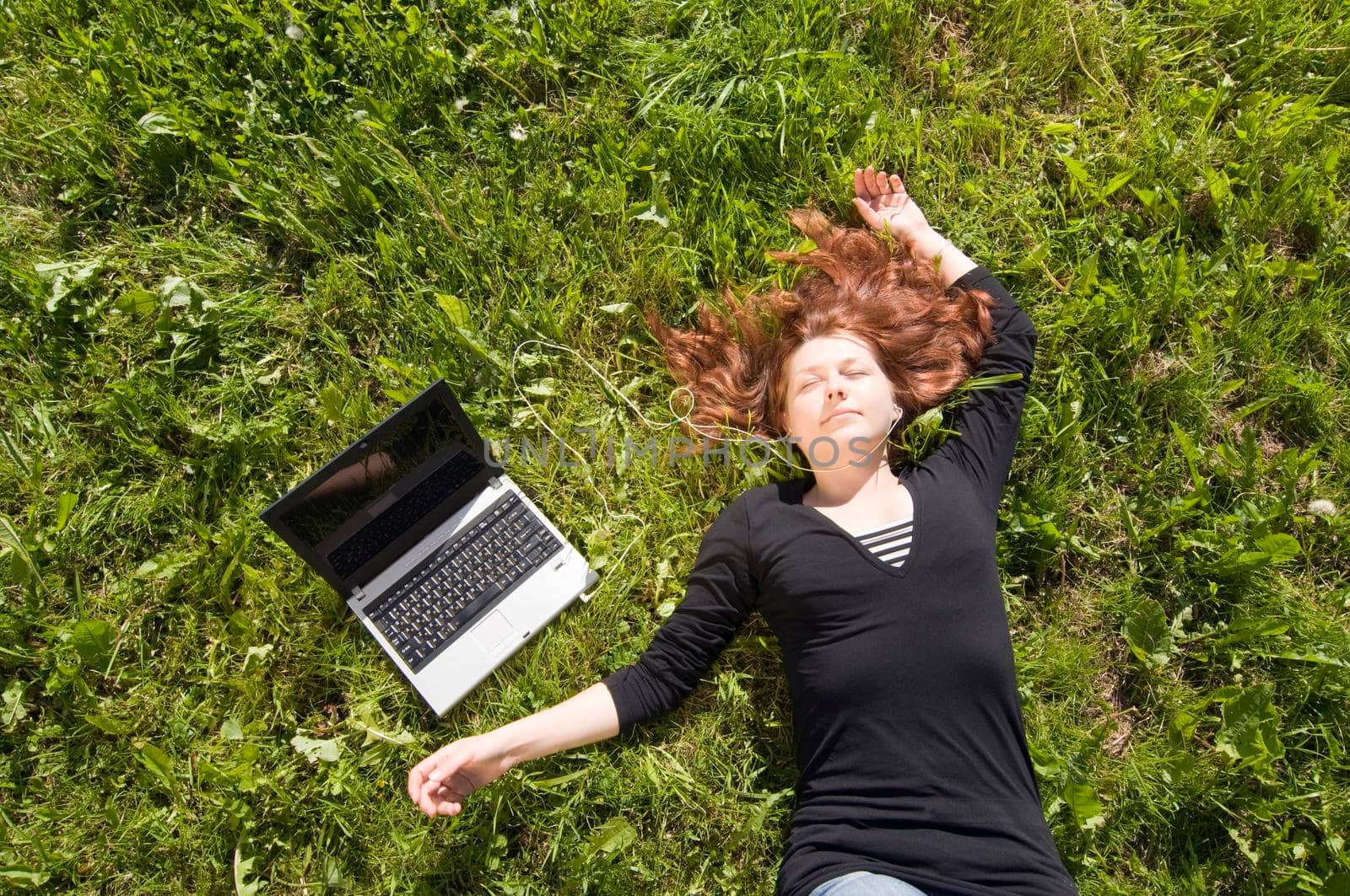 Beautiful girl lying on a grass and listening to the music from her laptop