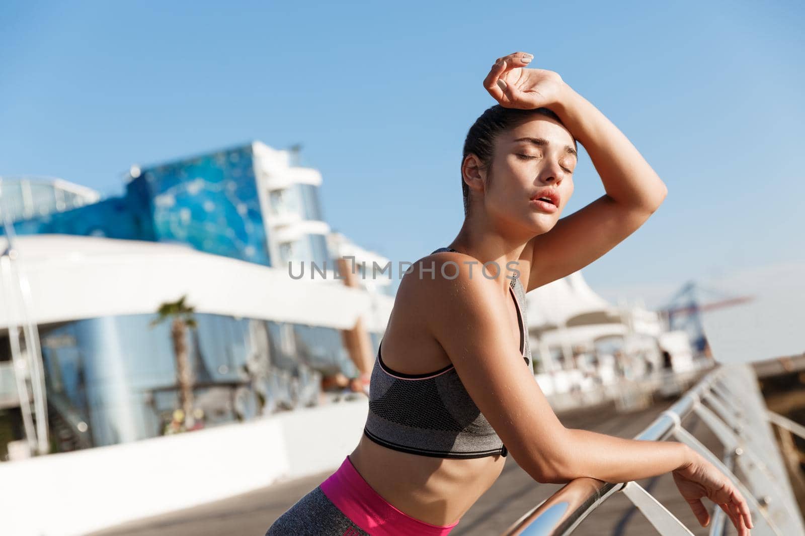 Close-up of attractive young fitness woman leaning on handrail, wiping sweat off forehead after workout, finish jogging on the seaside promenade.