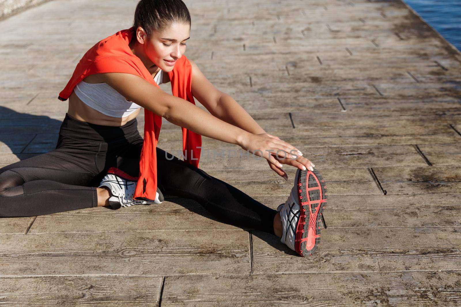 Outdoor shot of fit and healthy sportswoman stretching her legs and workout along seaside promenade by Benzoix