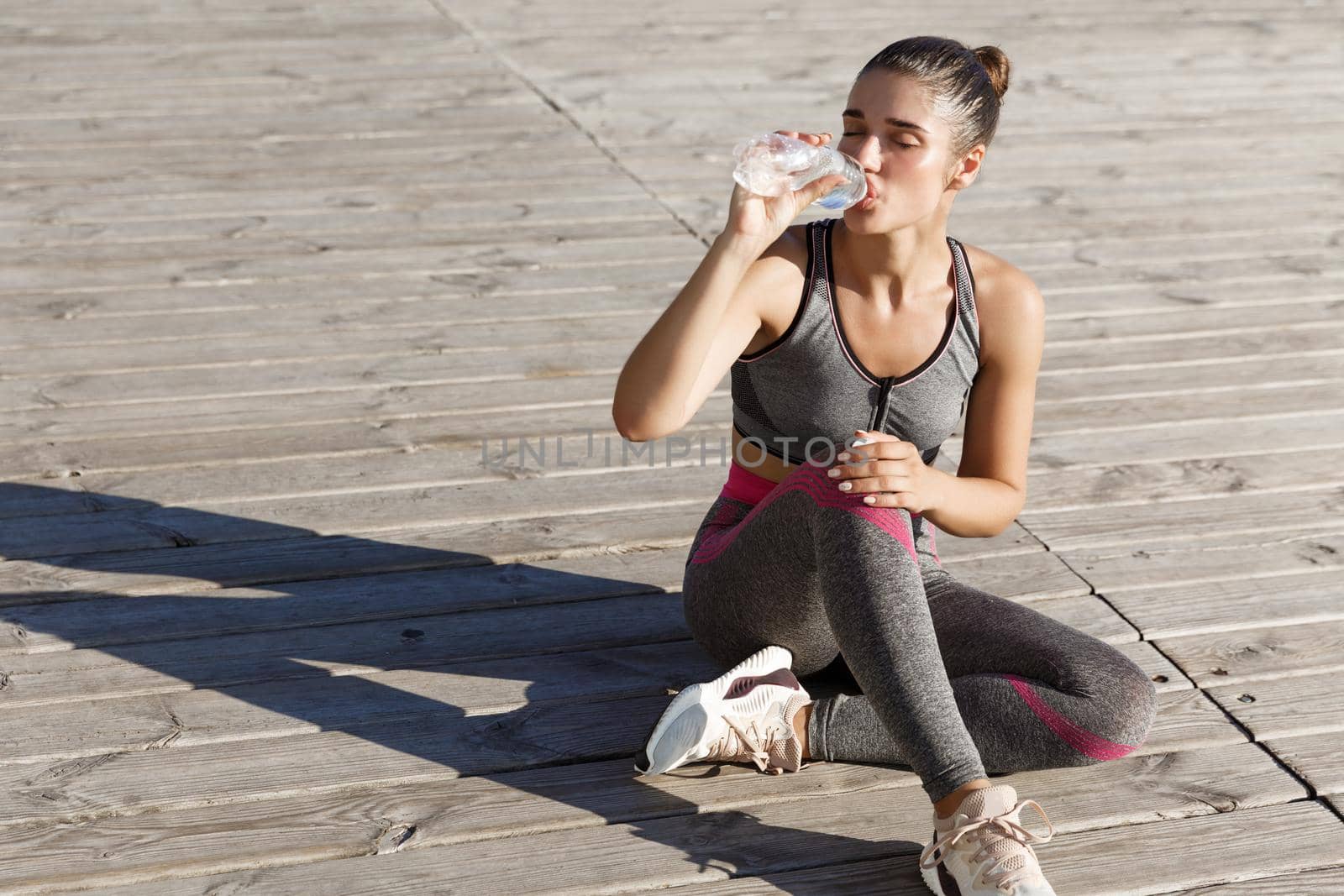 Close-up of attractive sportswoman taking a break after workout, sitting at the seaside and drinking water, looking pleased.