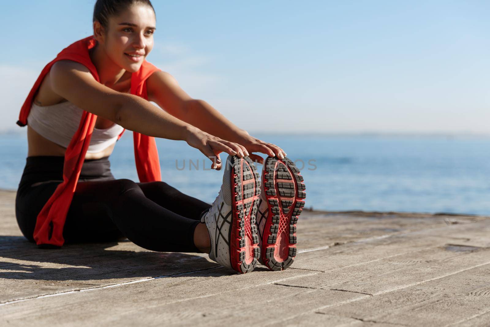 Outdoor shot of smiling confident sportswoman workout near sea. Fitness woman sitting on wooden pier and stretching legs by Benzoix