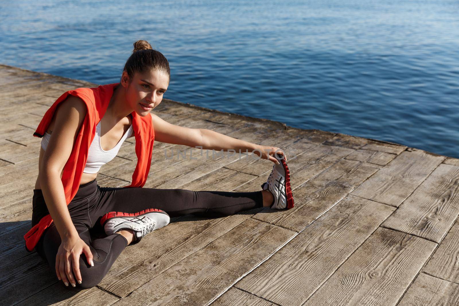 Outdoor shot of young sportswoman stretching legs while sitting on pier, having workout training on fresh air near the sea.