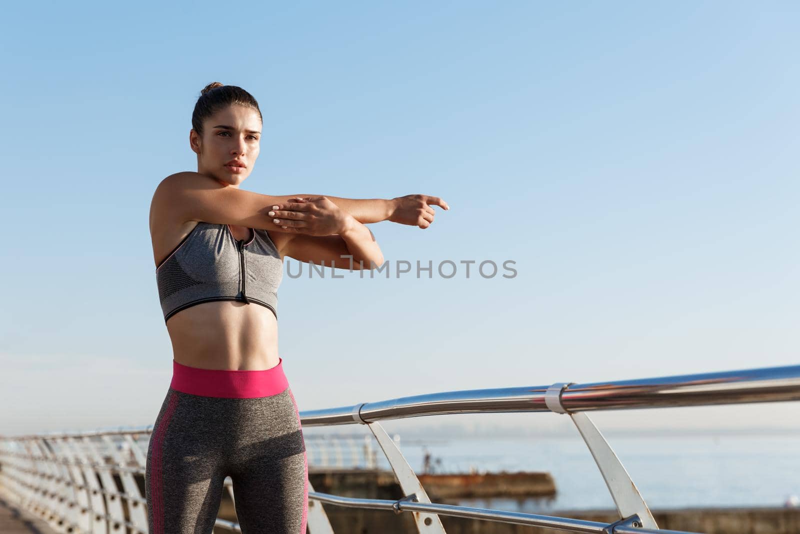 Young serious female athlete in sportswear stretching her body before jogging workout. Sportswoman standing on a pier and doing warm-up exercises before running.