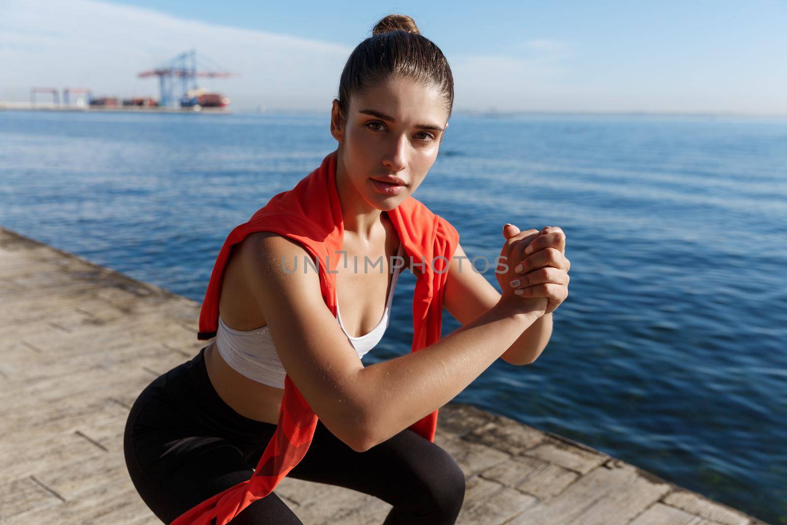 Close-up of confident fitness woman doing squats exercises near the sea, looking at camera during workout by Benzoix