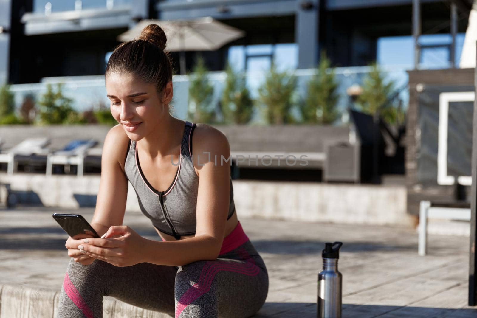 Outdoor shot of young female athlete sitting on a bench and looking at smartphone, resting after workout by Benzoix