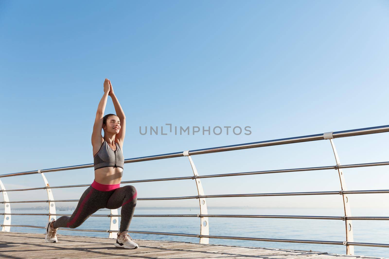 Outdoor shot of attractive sportswoman stretching and working out on the seaside promenade, looking at the sea and training by Benzoix