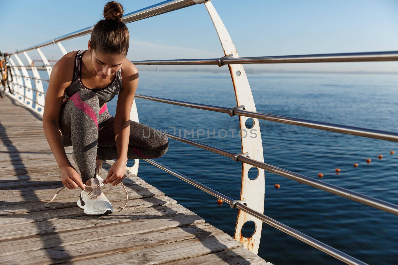 Outdoor shot of attractive sportswoman tying shoelaces on a pier, jogging at seaside by Benzoix