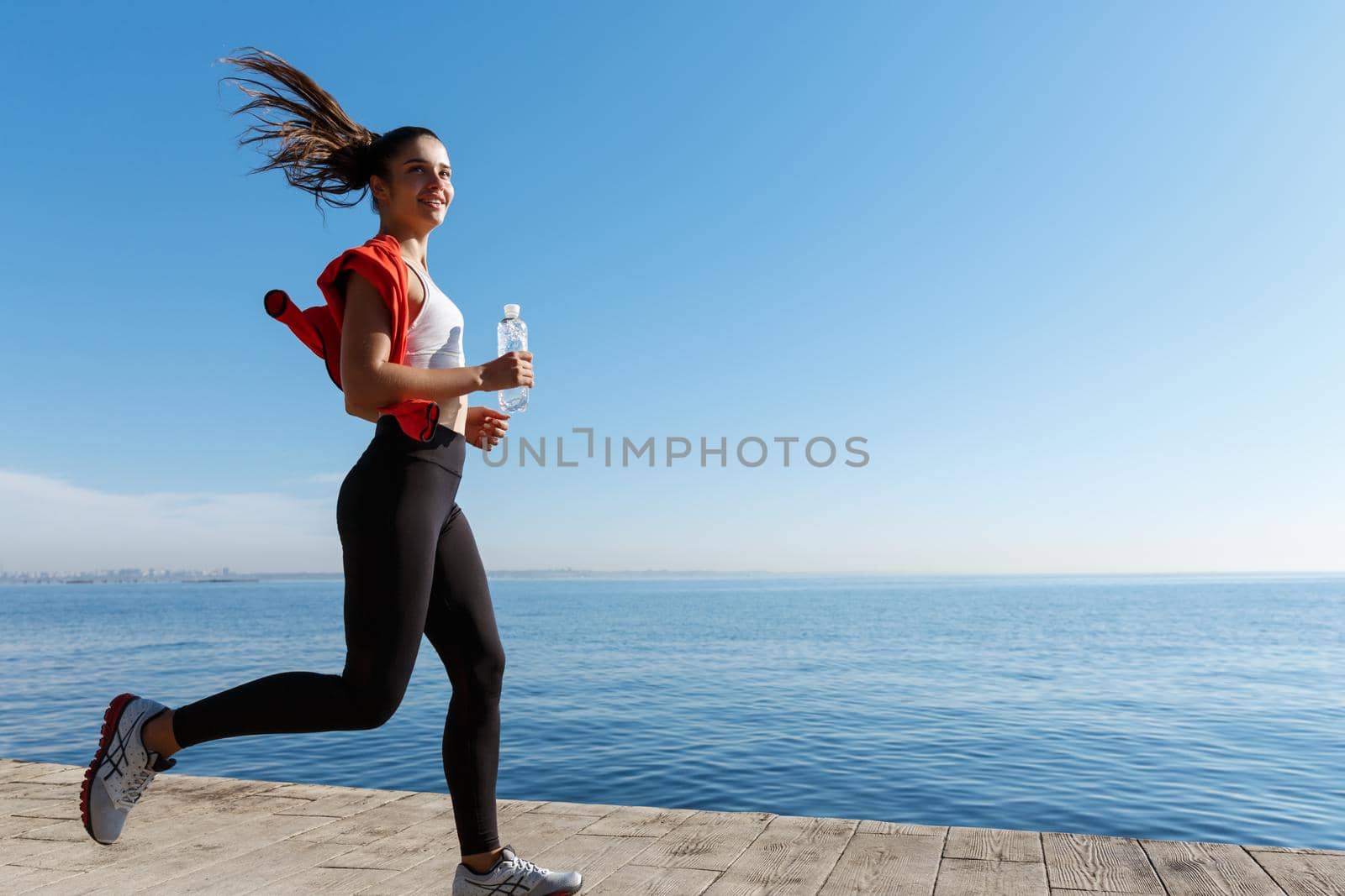 Side view of attractive and healthy fitness woman running along the seaside promenade. Girl jogging in the morning and smiling by Benzoix