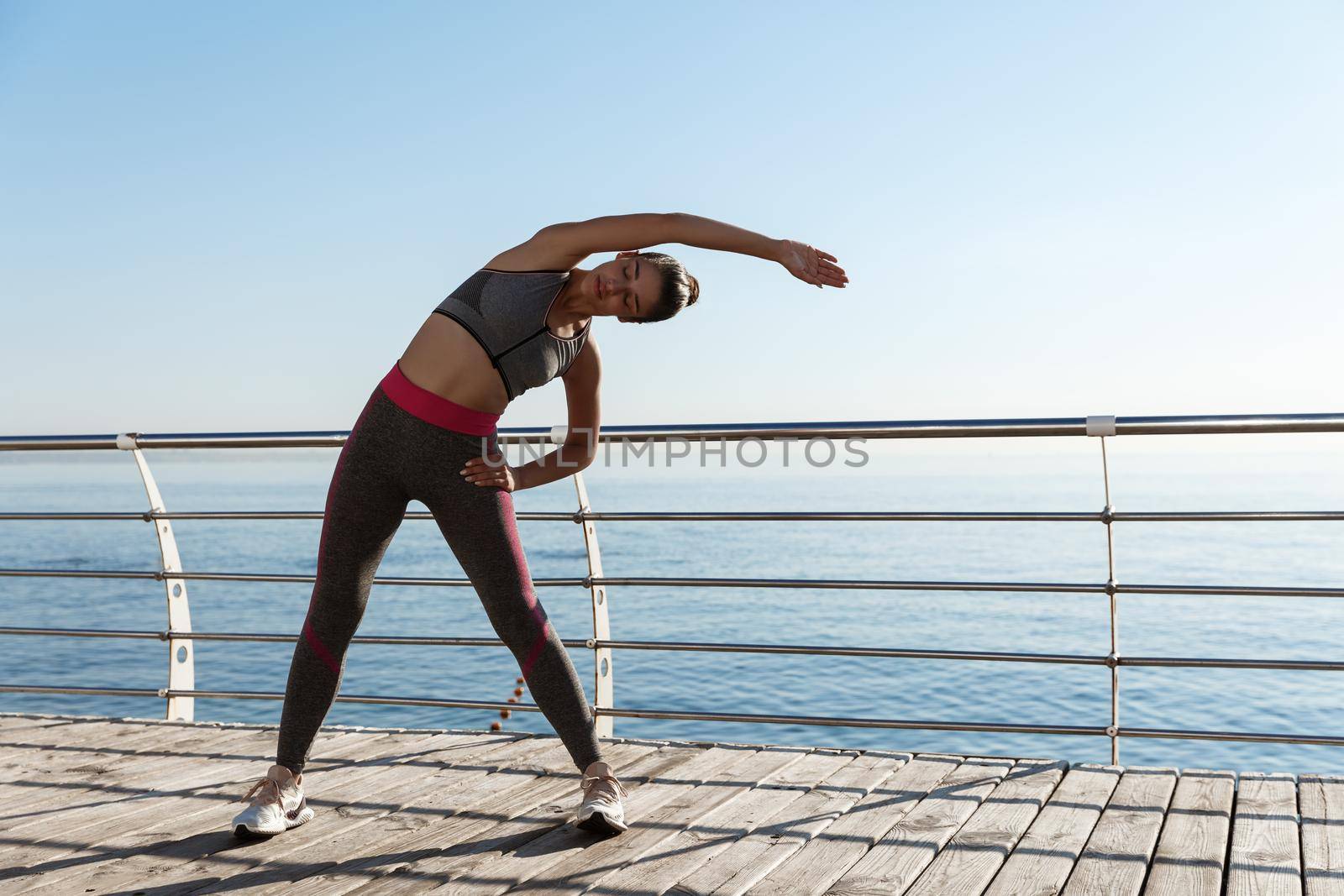 Outdoor shot of attractive and fit sportswoman workout on a pier. Girl stretching and training alone at the seaside.