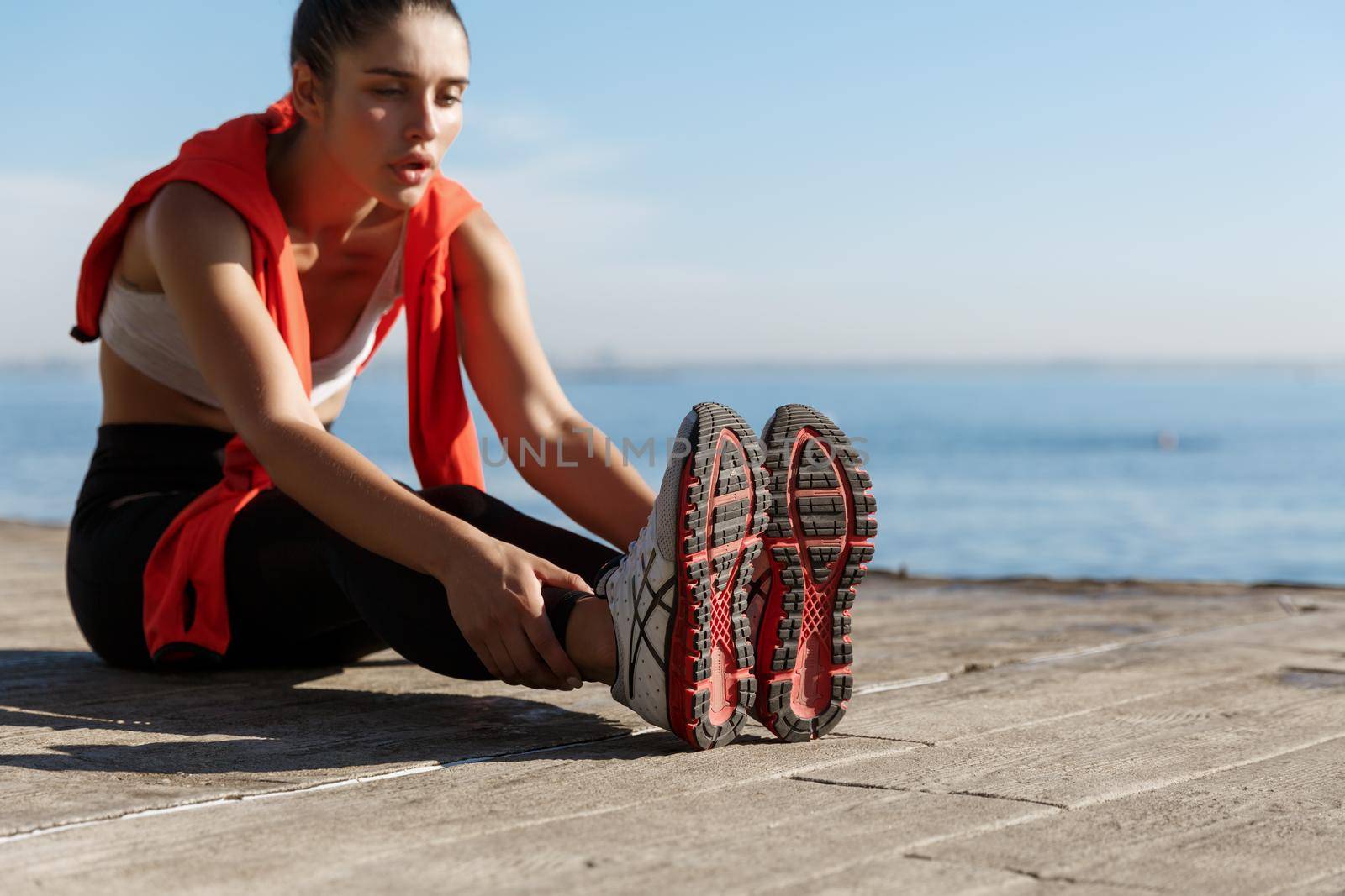 Outdoor shot of smiling confident sportswoman workout near sea. Fitness woman sitting on wooden pier and stretching legs.