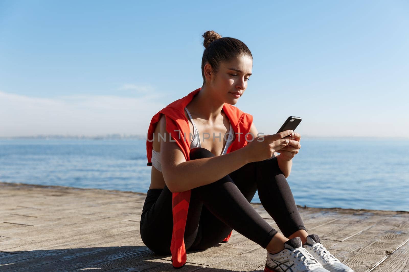 Outdoor shot of attractive sportswoman having a break near sea, sitting on wooden pier and using mobile phone.