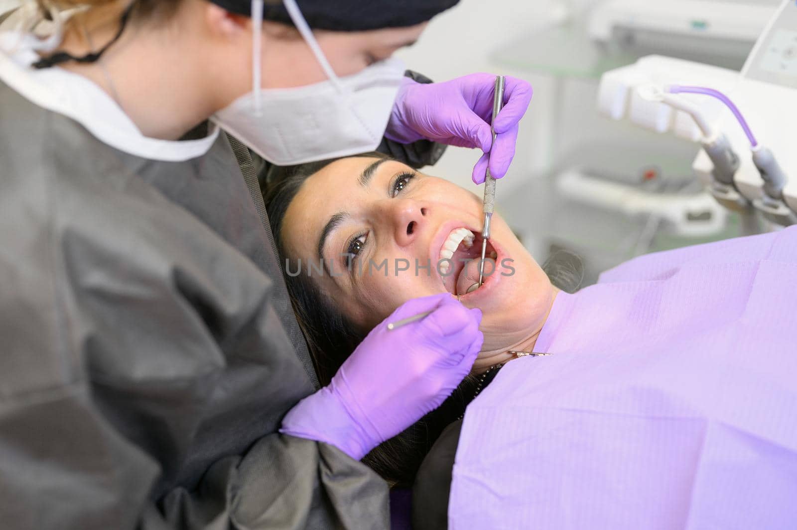 Young female patient visiting dentist office. Beautiful woman sitting at dental chair with open mouth during oral checkup while doctor working at teeth by HERRAEZ