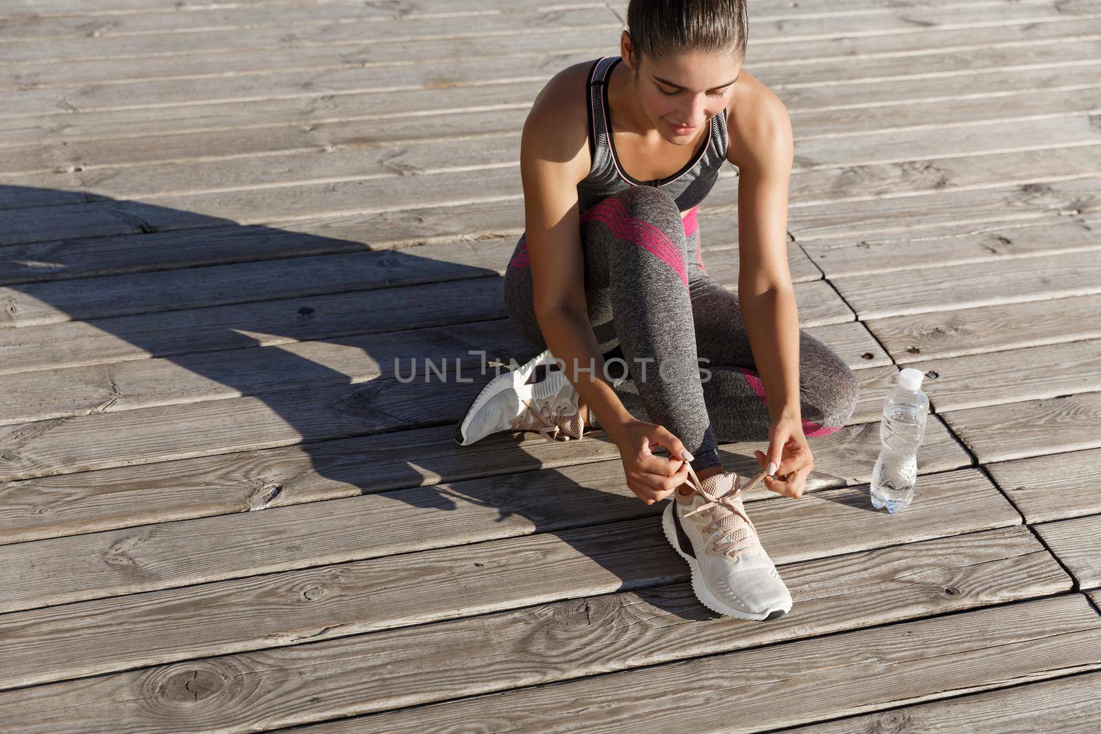 Outdoor shot of young fitness woman tying shoelaces and drinking water during workout.