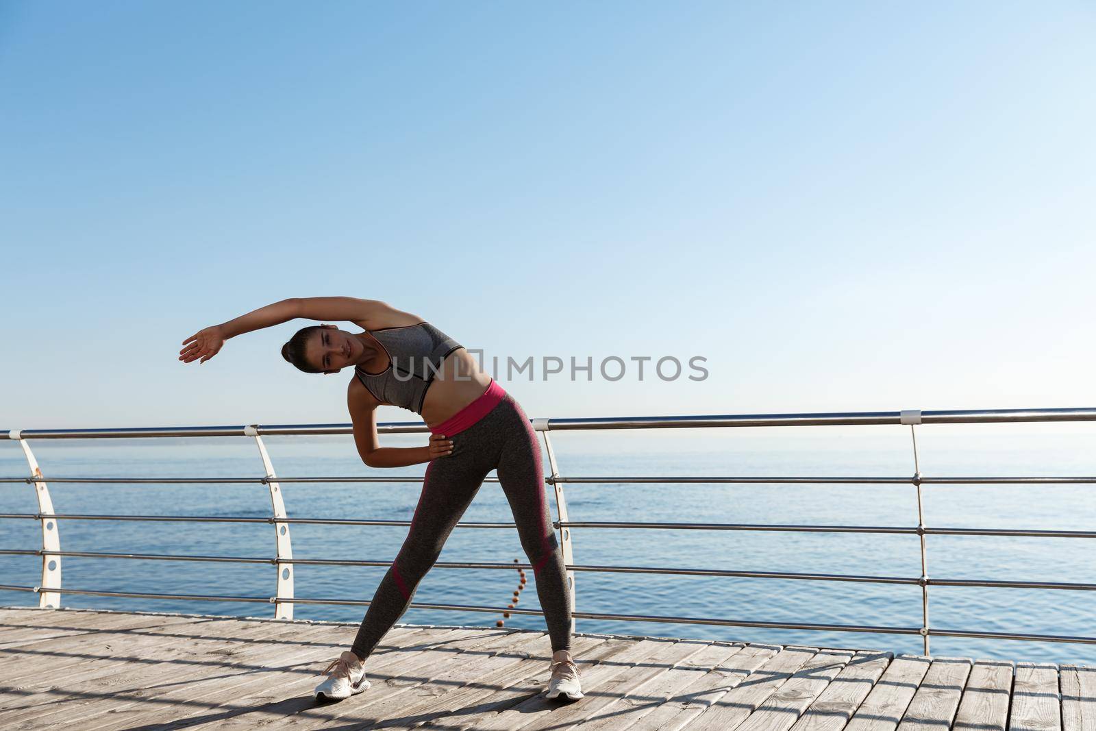 Outdoor shot of young sportswoman stretching and training on a pier. Female athlete workout near the sea by Benzoix