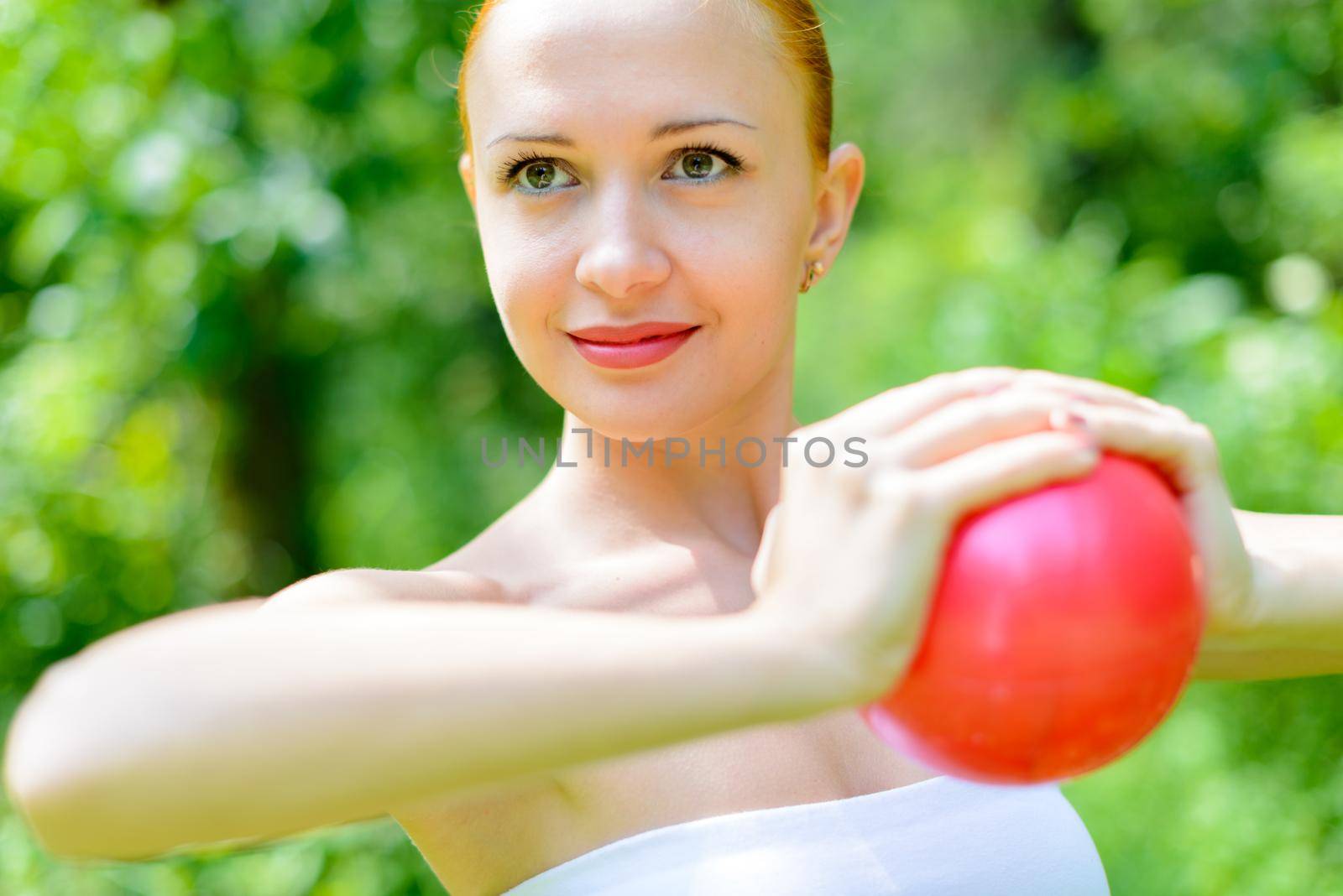 Young red female fitness instructor exercising with small ball in green park