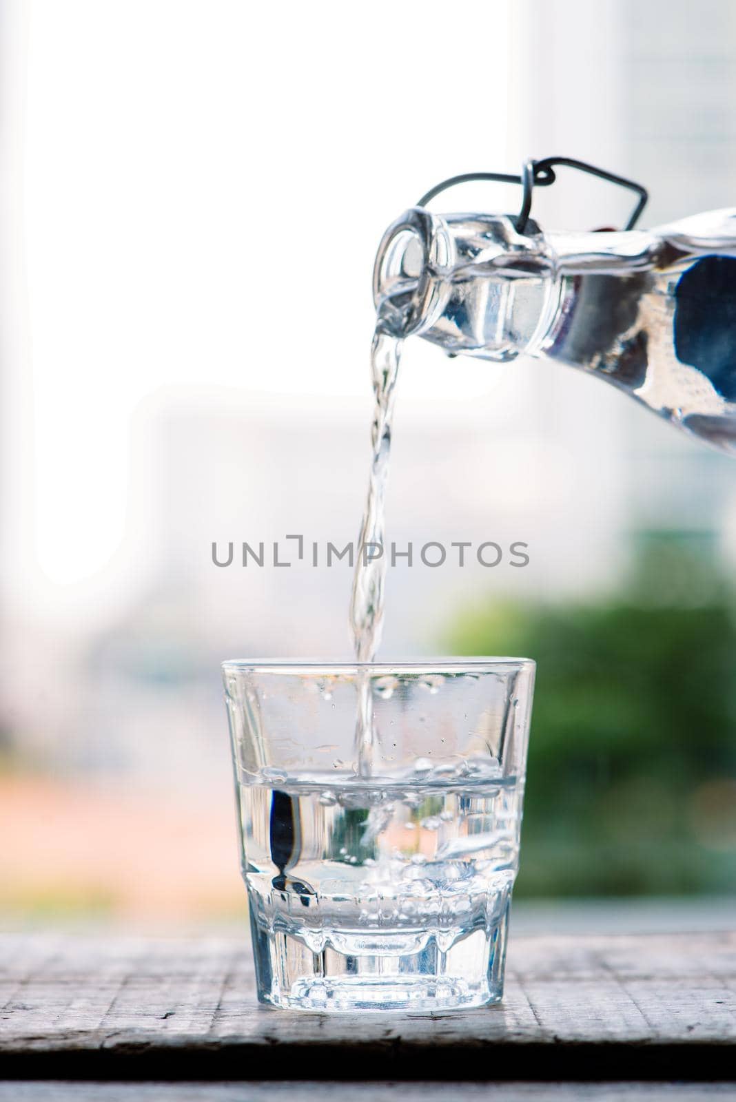 Clean drinking water is poured from a jug into a round glass cup on a wooden table and a light green napkin close-up macro on a green nature outdoors background .