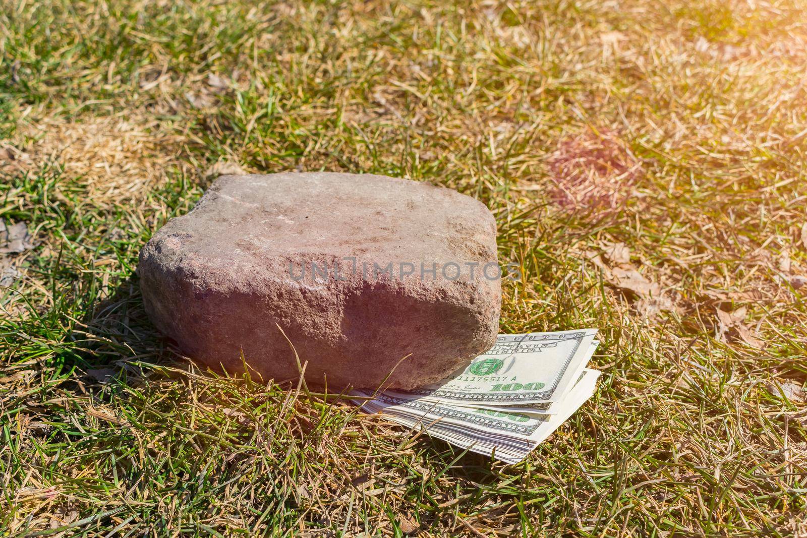 A large bundle of money lies on the ground, pressed against the grass by a stone