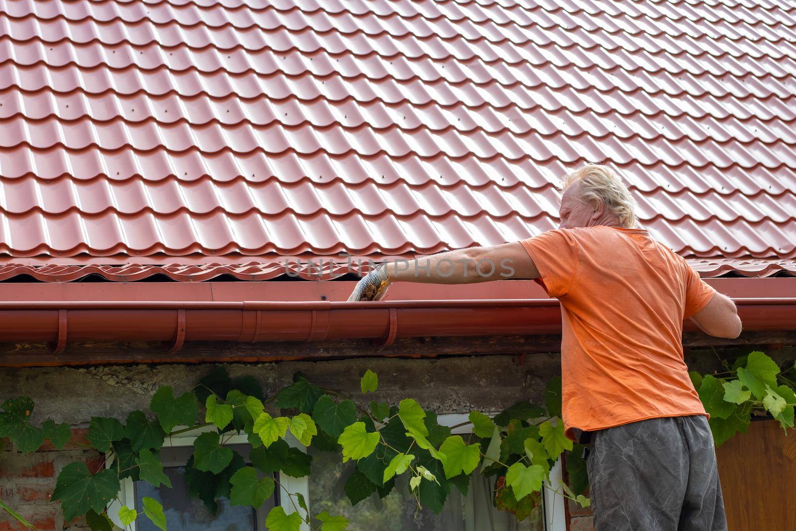 A man cleans out debris and leaves from the gutter system on the roof of his house by levnat09