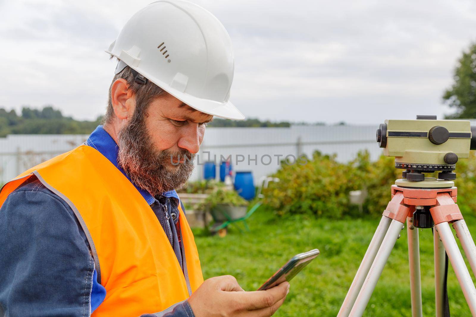 A civil engineer with an optical level looks into a mobile phone. A bearded man is looking for information on his mobile phone.
