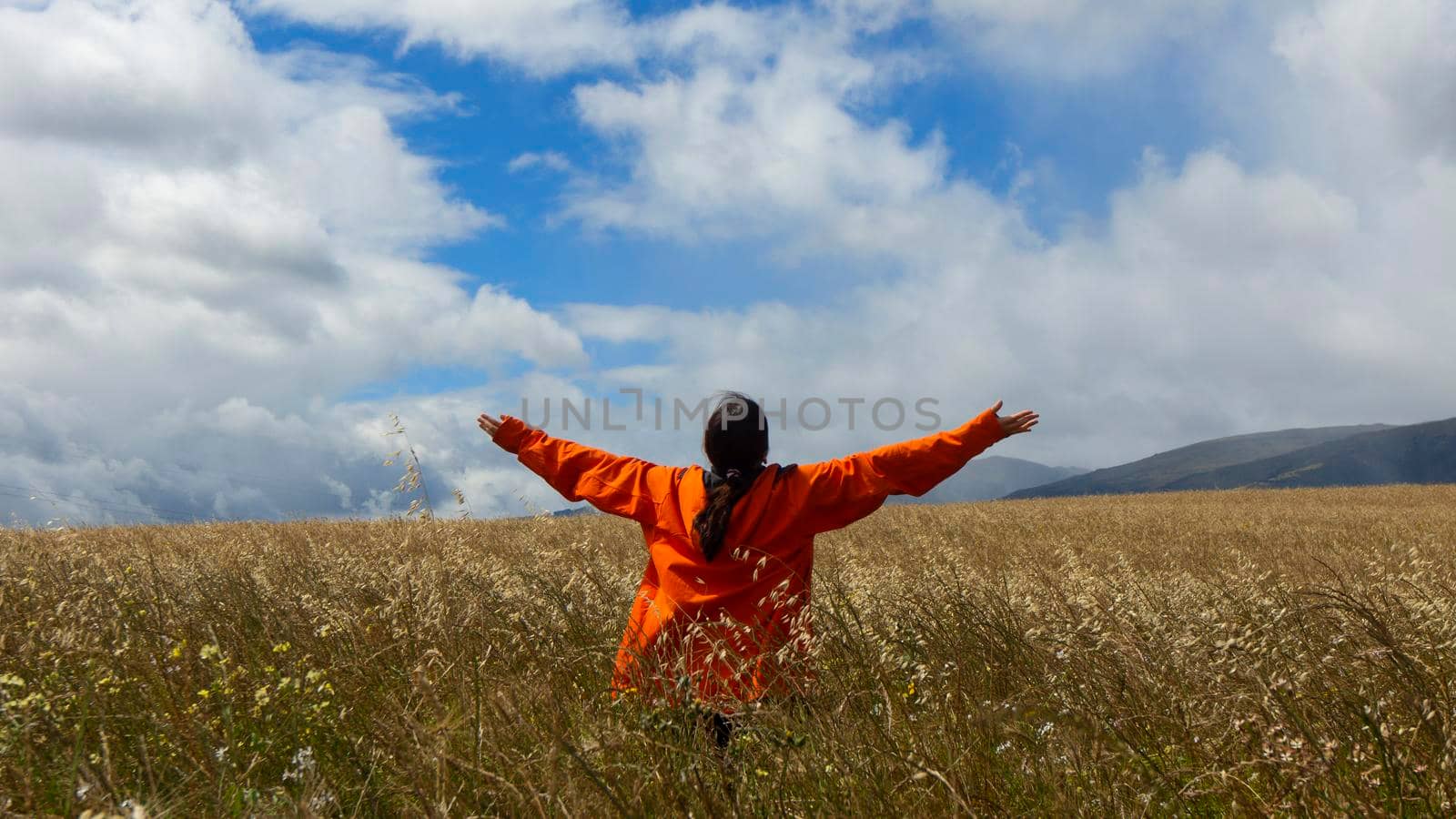 Young woman from behind in orange jacket with raised arms looking up at the sky in the middle of a wheat field on a cloudy day with blue sky and mountains in the background