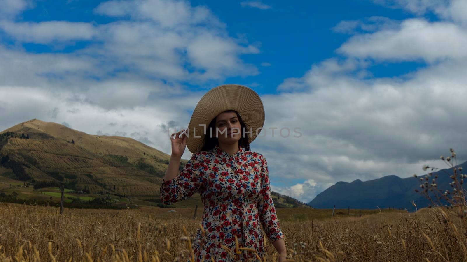 Happy young woman in floral dress holding her hat with her right hand walking alone in the middle of a wheat field seen from the front on a cloudy day with blue sky by alejomiranda
