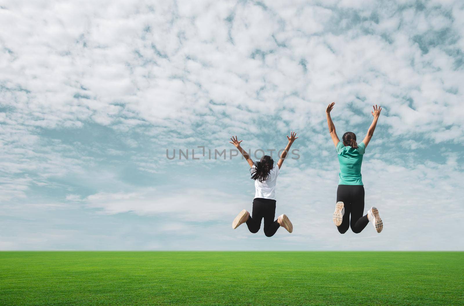 Mom and girl jumping in green field against blue sky. by thanumporn