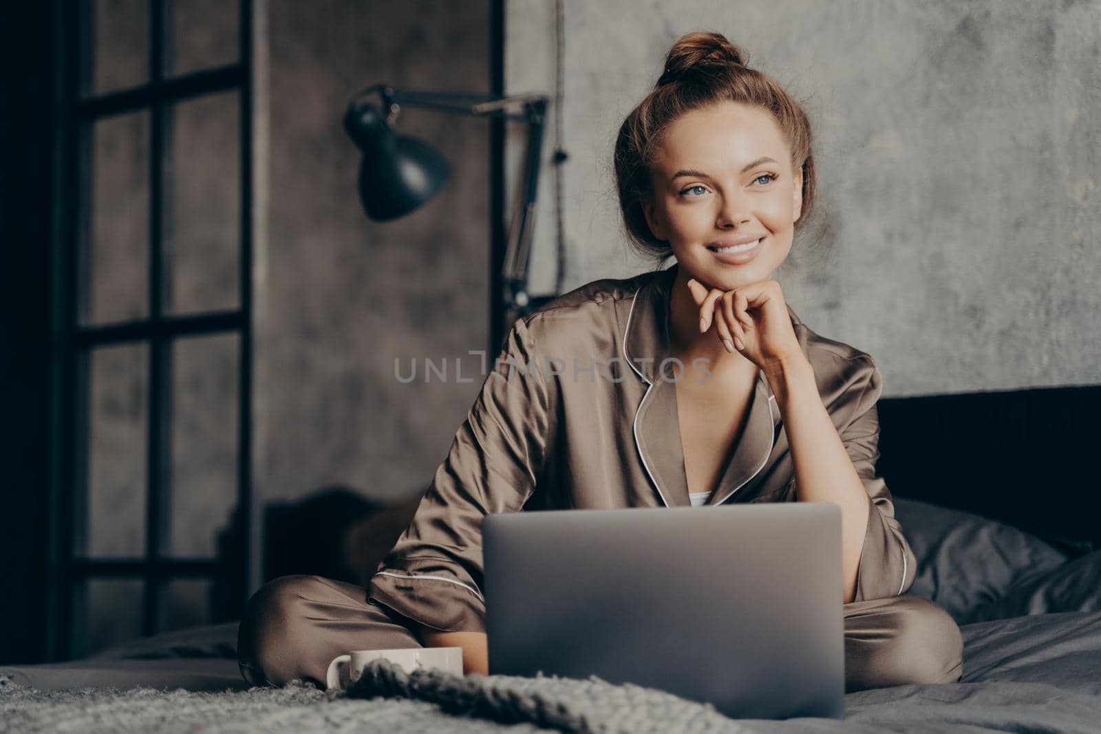 Portrait of beautiful thoughtfully smiling businesswoman in satin pajama working remotely from home in morning, sitting on bed with laptop checking emails from work and enjoying time after waking up