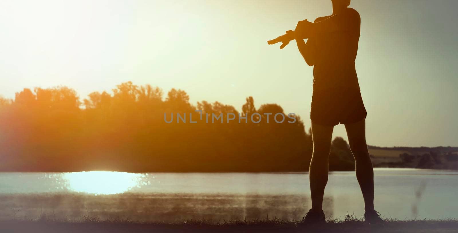 A young man trains outdoors against the backdrop of a beautiful lake at sunset, the athlete warms up, does exercises on the arms and shoulders.