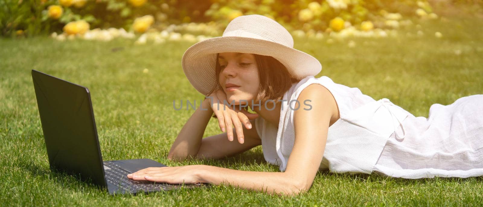 A young girl in a white dress is working on a laptop, texting on the Internet on her device, freelancing, studying, resting on a meadow in a park, lying on the grass.