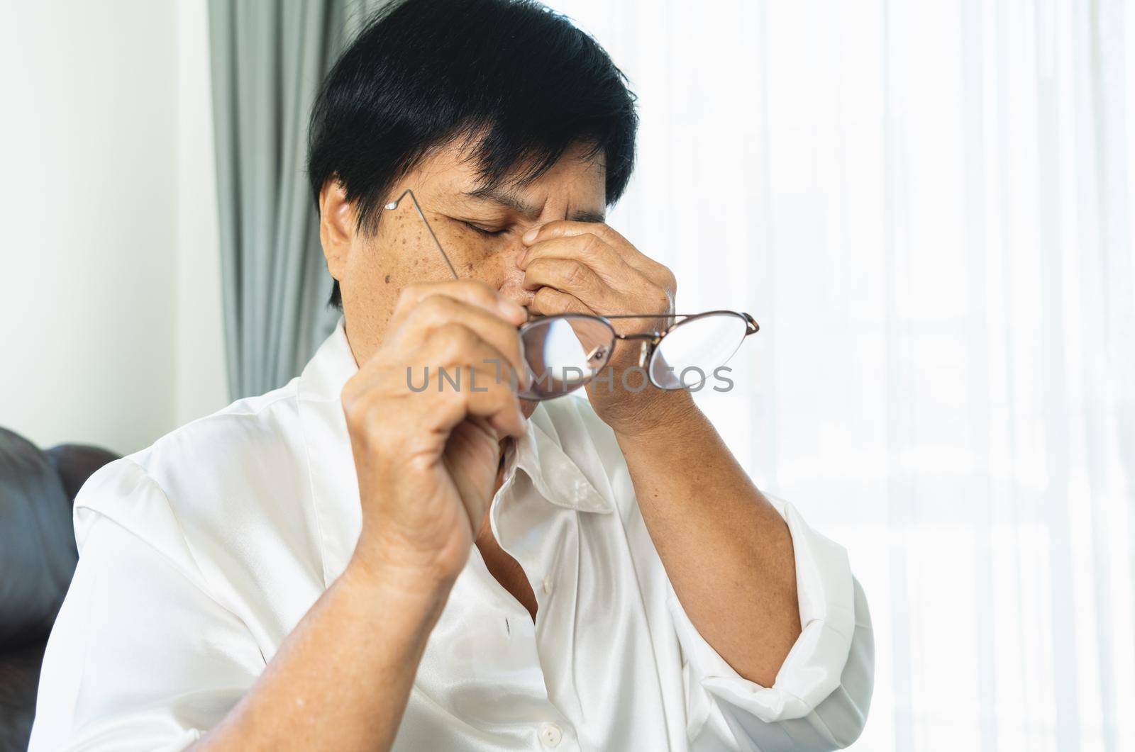 Tired old woman removing eyeglasses, massaging eyes after reading paper book. feeling discomfort because of long wearing glasses, suffering from eyes pain or headache