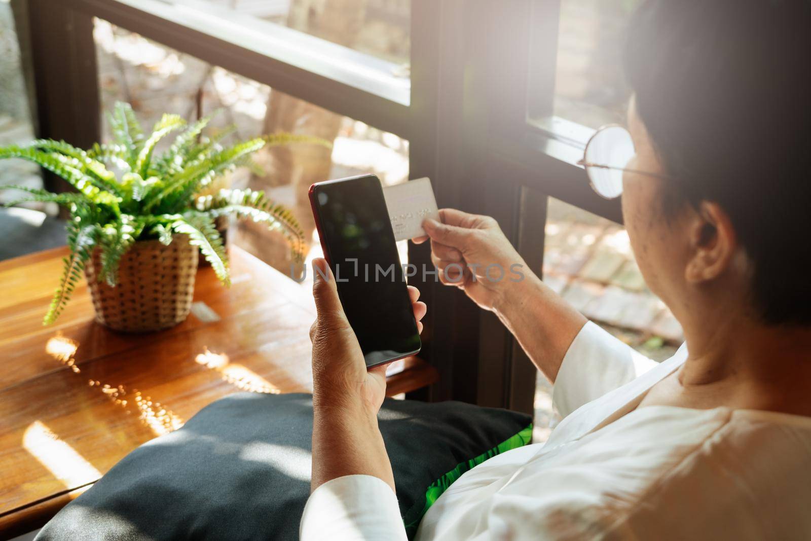 senior lady sitting in a chair and holding the credit card with smartphone, online shopping concept by psodaz