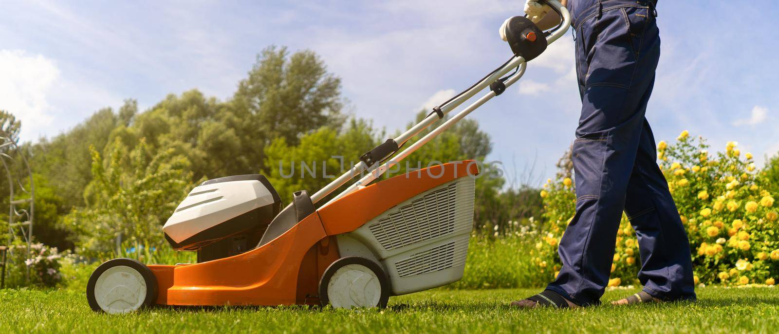 A young man in a straw hat is mowing a lawn with a lawn mower in his beautiful green floral summer garden. A professional gardener with a lawnmower cares for the grass in the backyard.