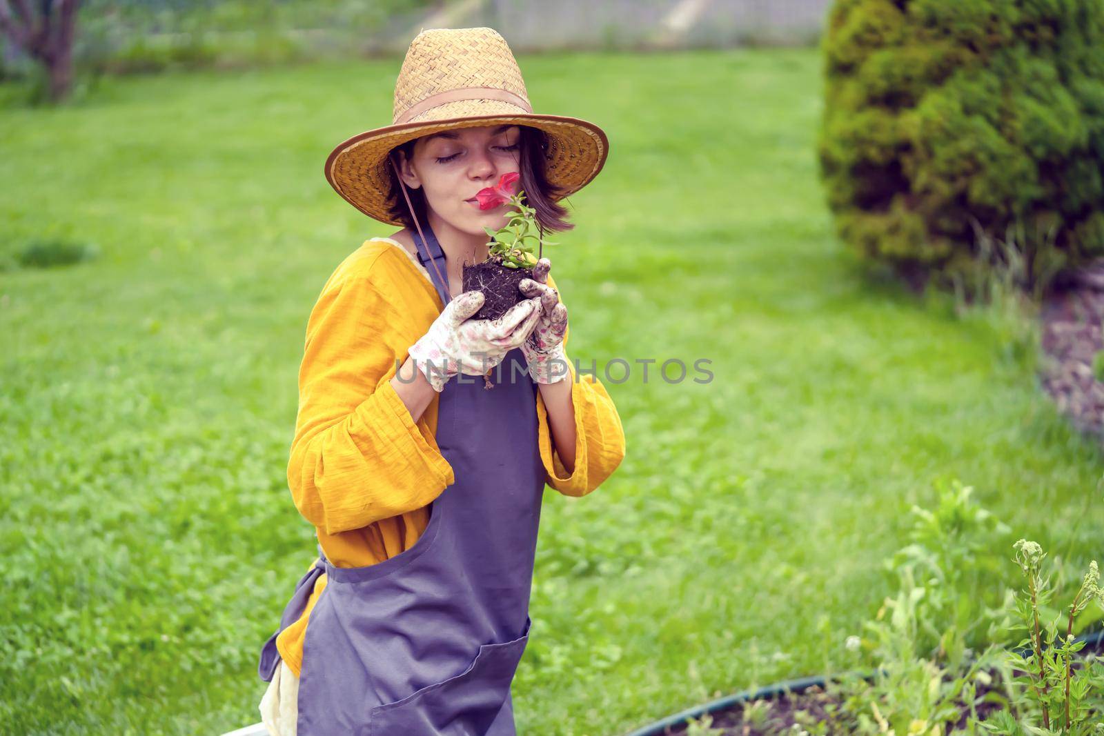 A young woman gardener in a straw hat and hands in gloves is holding a petunia flower in a peat pot, sniffing the flower and smiling before planting a seedling