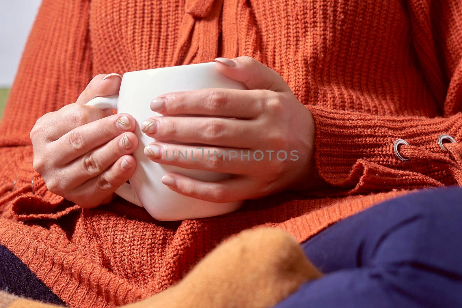 Autumn mood. Young female in orange woolen pullover drinking refreshing coffee in a cold autumnal morning. Woman in sweater holding in hands a cup of tea