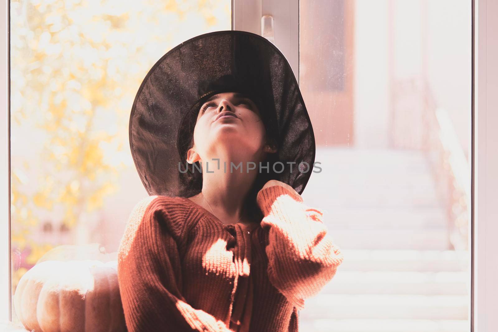 Halloween lady wearing witch cup. A woman in witch hat and Halloween pumpkin near the windows. Portrait of young female in orange sweater and traditional black witch cap