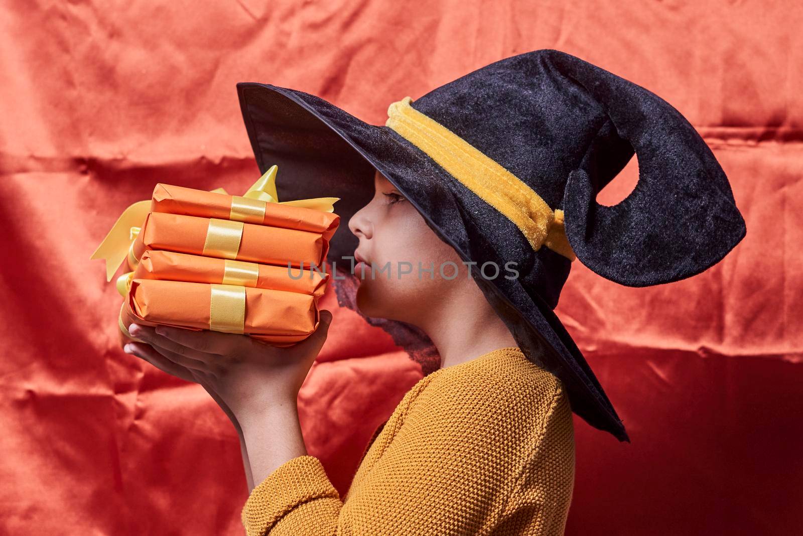A little kid in witch cap holding a pile of gift boxes. Halloween gifts in hands of a boy over orange background