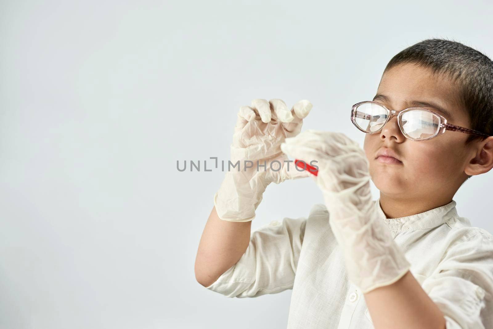 A portrait of a cute boy in protective gloves and glasses holding a piece of red plasticine in hands against the white background