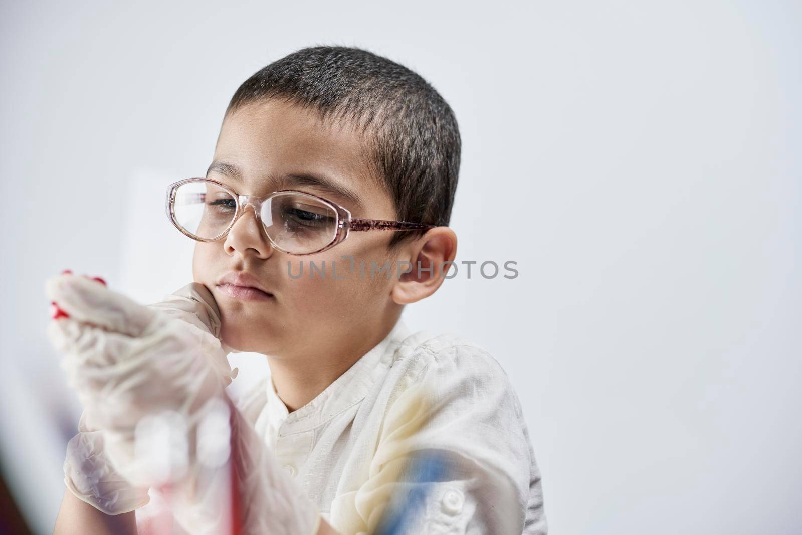 A portrait of a cute boy in protective gloves and glasses holding a piece of red plasticine in hands against the white background