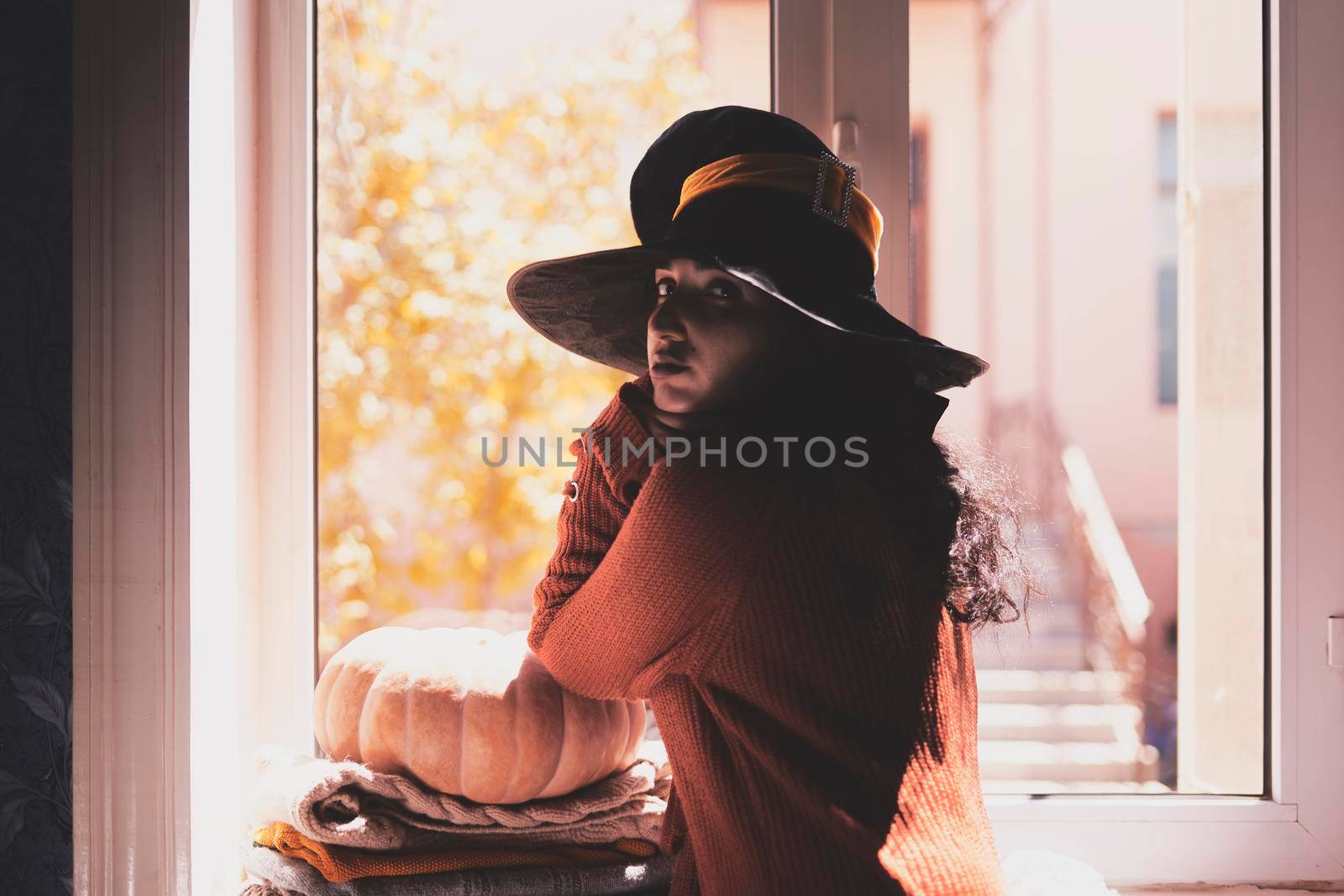 Halloween lady wearing witch cup. A woman in witch hat and Halloween pumpkin near the windows. Portrait of young female in orange sweater and traditional black witch cap