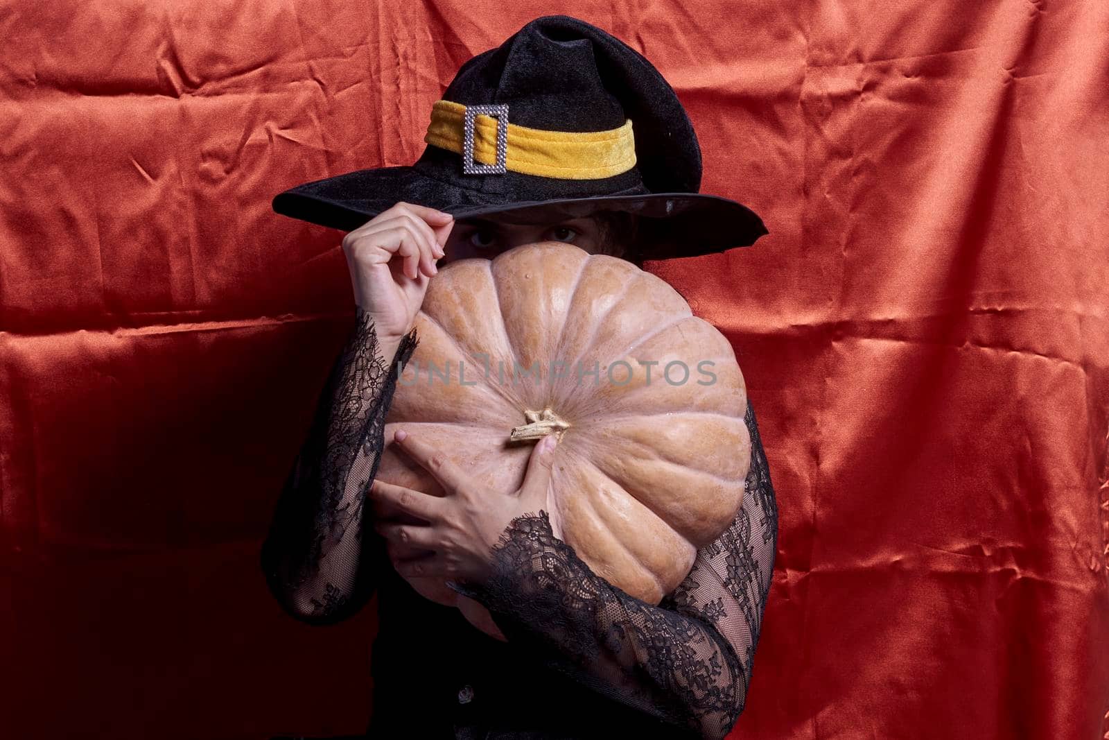 Halloween lady wearing witch cup and holding a big Halloween pumpkin. A woman in witch hat and Halloween pumpkin over orange background. Portrait of young female in traditional black witch cap