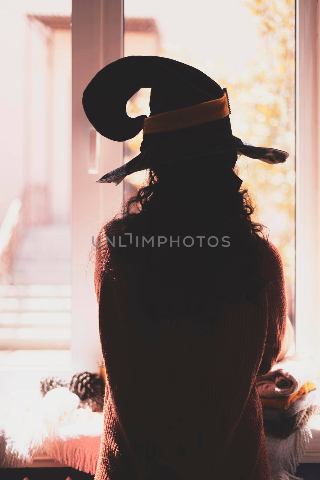 Halloween lady wearing witch cup. A woman in witch hat and Halloween pumpkin near the windows. Portrait of young female in orange sweater and traditional black witch cap