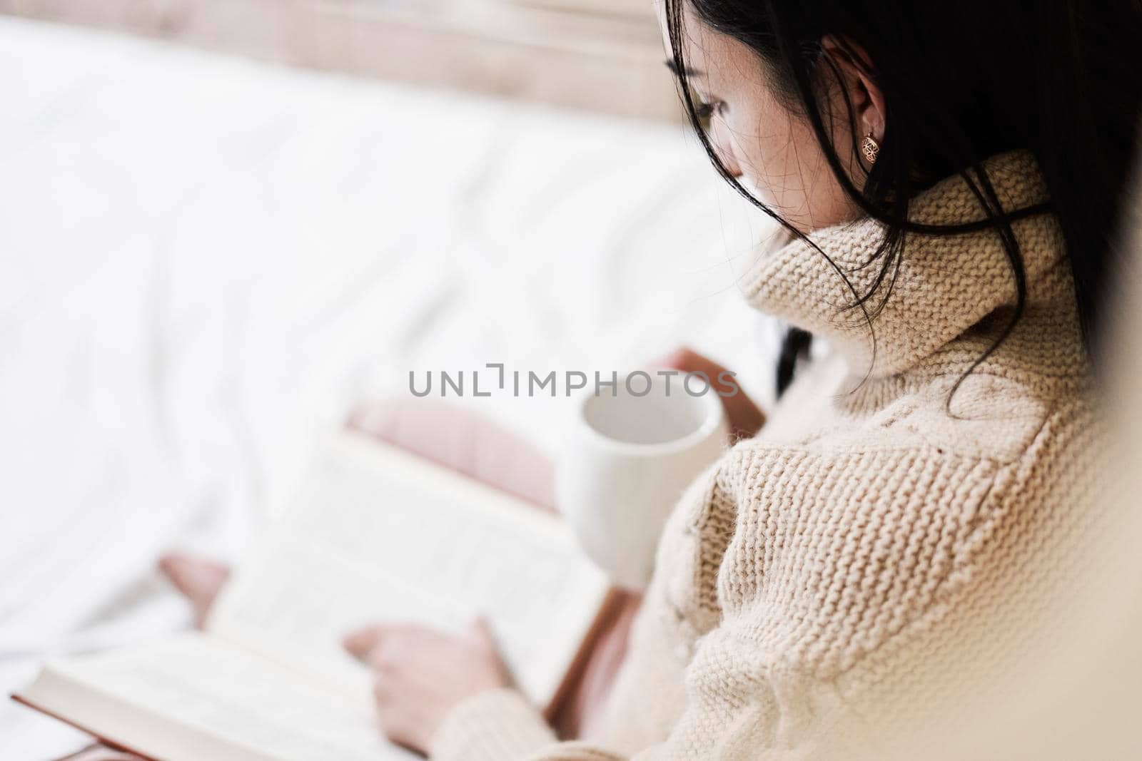 Reading in bed. Young woman drinking coffee and reading a book in the bed. Multi-racial female reads a book in the bed. Cozy autumn morning in the bedroom. Young female enjoys weekend