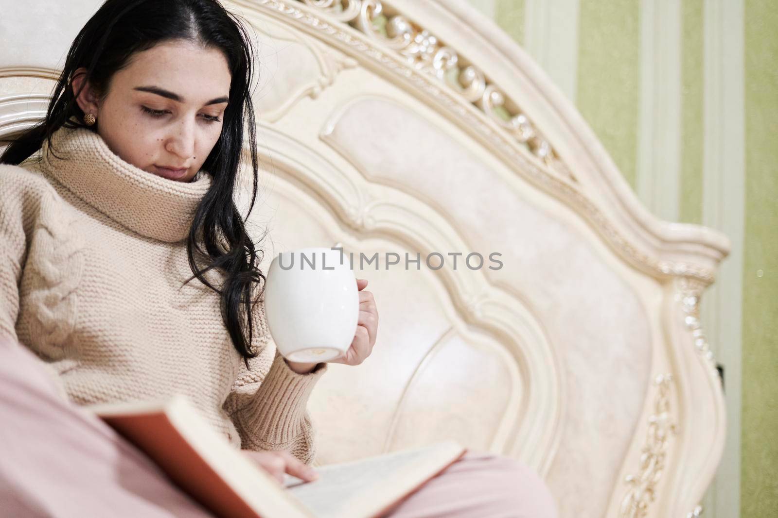 Reading in bed. Young woman drinking coffee and reading a book in the bed. Multi-racial female reads a book in the bed. Cozy autumn morning in the bedroom. Young female enjoys weekend