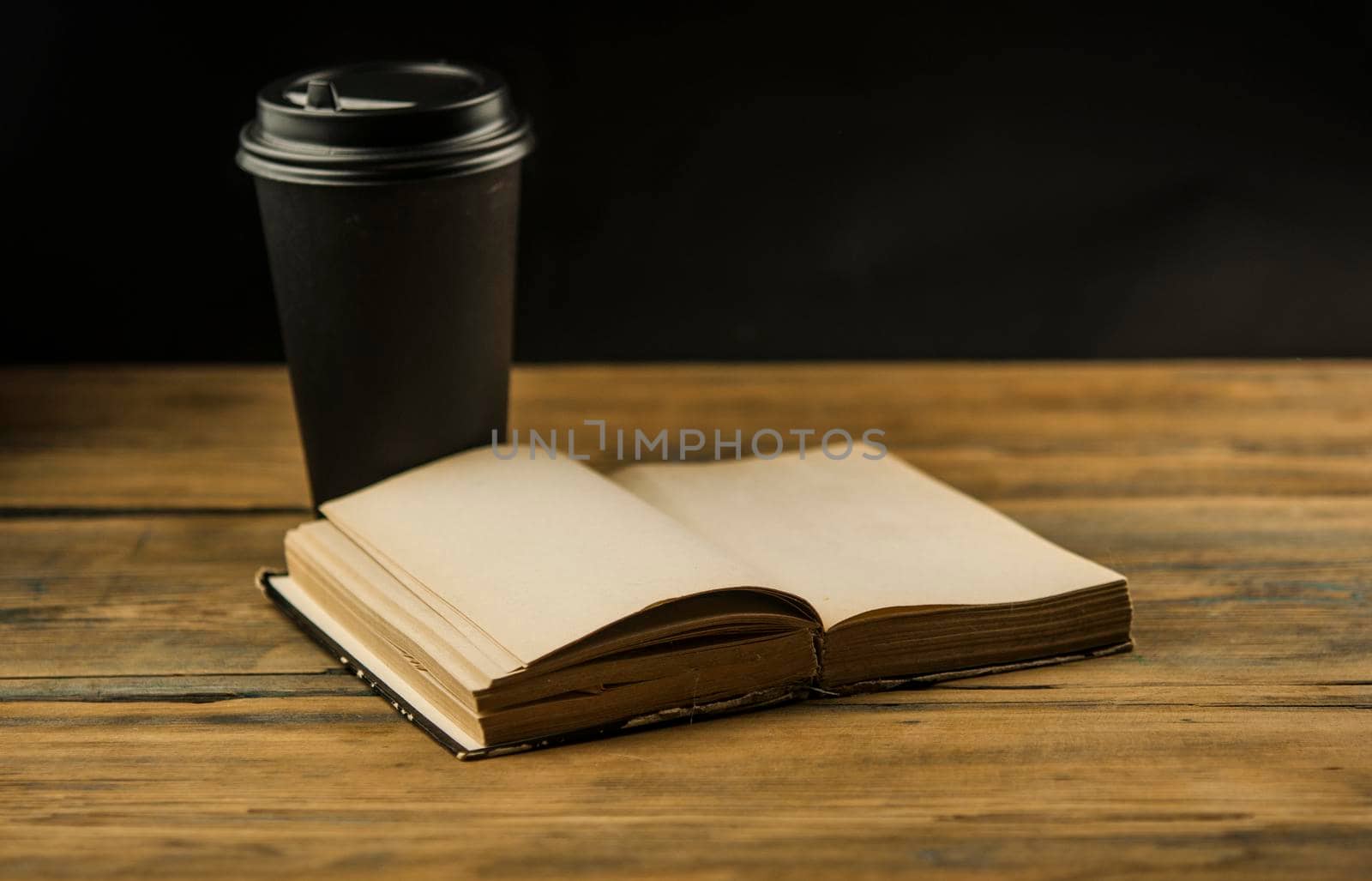 Open book and a cup of coffee in a disposable paper cup on a wooden table