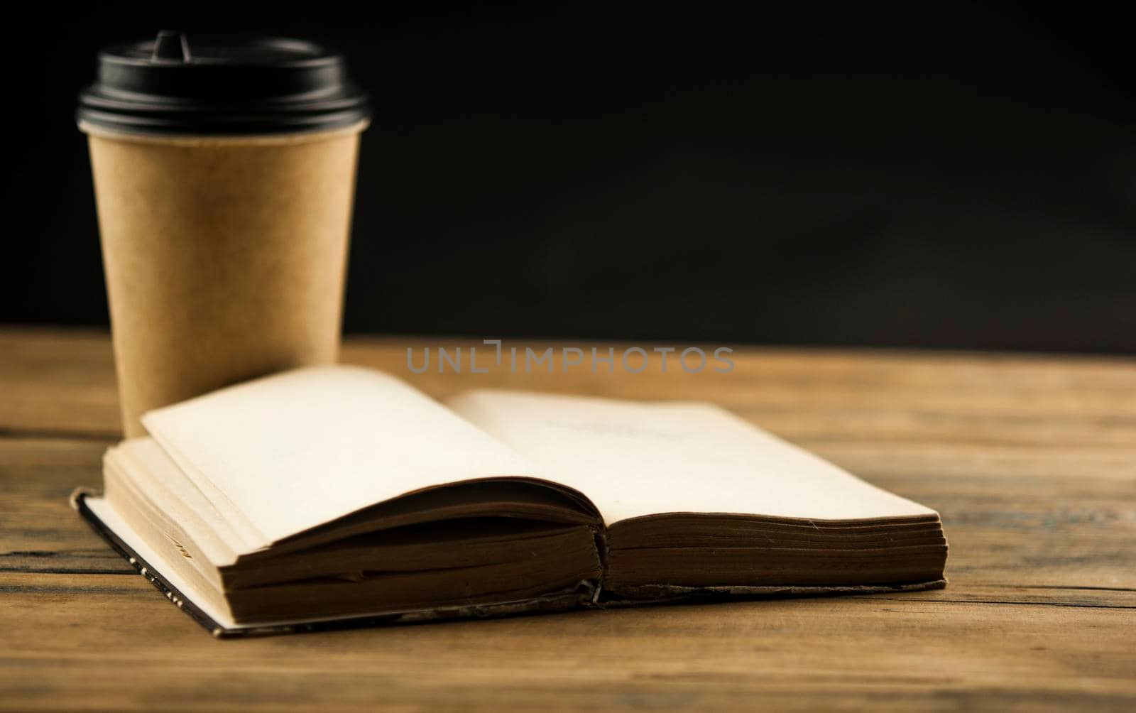 Open book and a cup of coffee in a disposable paper cup on a wooden table by inxti