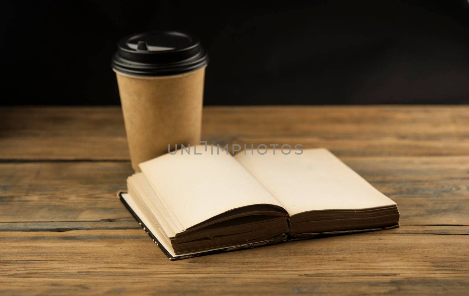 Open book and a cup of coffee in a disposable paper cup on a wooden table by inxti