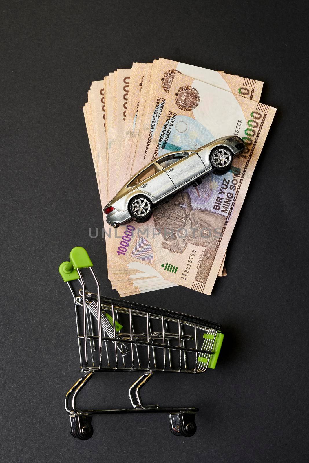 Buy auto insurance in Uzbekistan. Shopping cart, Uzbek sums and car toy. Concept of auto vehicle insuarance in Uzbekistan. Pile of 100000 sums and modern automobile toy