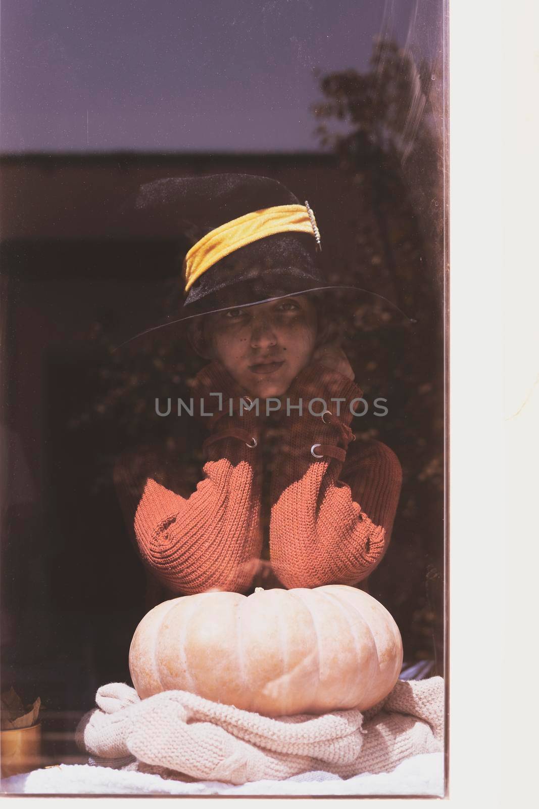 Halloween lady wearing witch cup. A woman in witch hat and Halloween pumpkin near the windows. Portrait of young female in orange sweater and traditional black witch cap